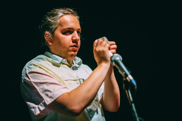 Robert Howat stands on a dark stage. He has his hair tied back and is wearing a light, short-sleeved shirt. In his hands, he holds a small, folded piece of paper. A microphone stands in front of him.