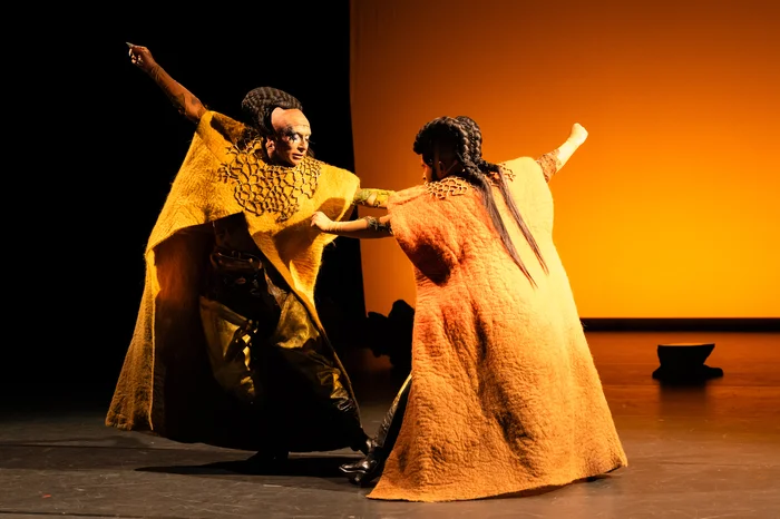 Zwei Performer*innen in großen, farbenfrohen Umhängen tanzen zusamenn, die Arme ausgebreitet. Die Bühne ist in warmes, goldenes Licht getaucht, das eine kraftvolle, fast rituelle Atmosphäre erzeugt