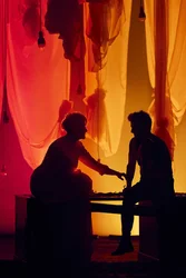 Zwei Personen sitzen sich als Silhouetten gegenüber und berühren sich leicht an den Händen; im Hintergrund hängen Stoffbahnen und Glühbirnen, in warmem Rot- und Orangeton ausgeleuchtet.