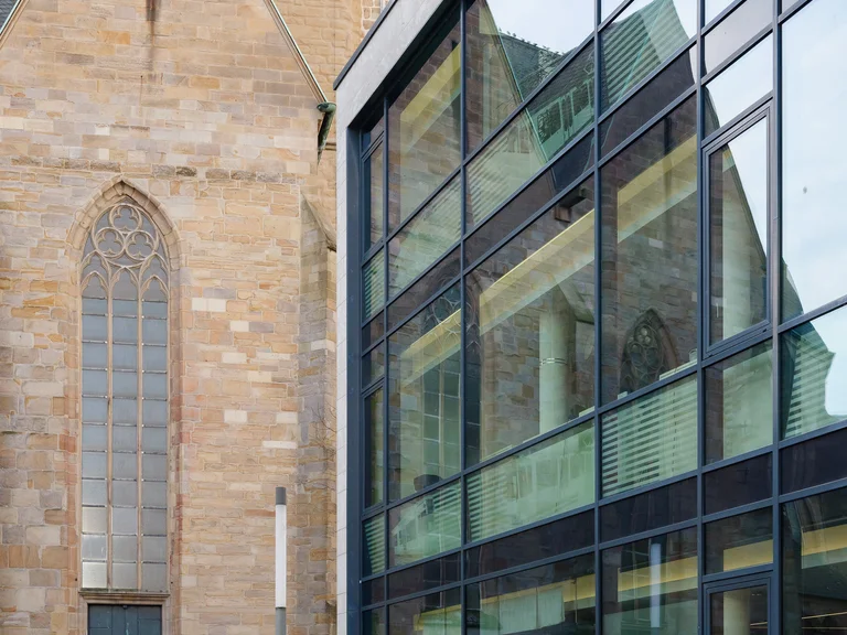 Eine historische Kirche aus hellem Stein steht unmittelbar neben einem modernen Gebäude mit großer Glasfassade.