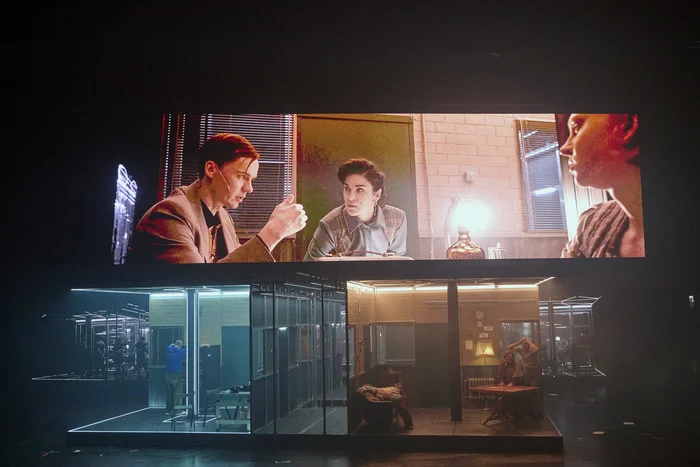 Stage scene with several actors in a glass-walled room; above them a large video projection showing a close-up of three characters.