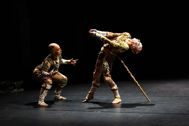 Two performers in elaborate, nature-inspired costumes move expressively on stage. The scene depicts a dance with powerful body language.