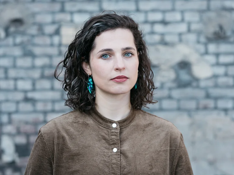 Eine junge Frau mit offenem Haar. Sie steht vor einer grauen Backsteinwand und blickt direkt in die Kamera.