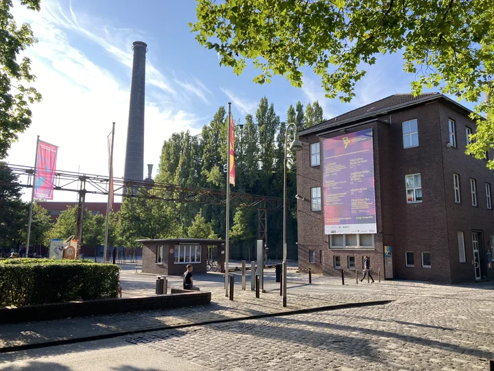 Eingang zum Landschaftspark Duisburg-Nord: Es sind alte Gebäude des ehemaligen Stahlwerks zu sehen. Am Rand gibt es Bäume, die Sonne scheint.