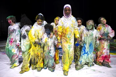 Eine Gruppe von Kindern in weißen Schutzanzügen mit bunten Farbflecken steht auf einer Bühne. Die Kinder sind lachend, spielerisch oder nachdenklich in verschiedene Richtungen blickend dargestellt.