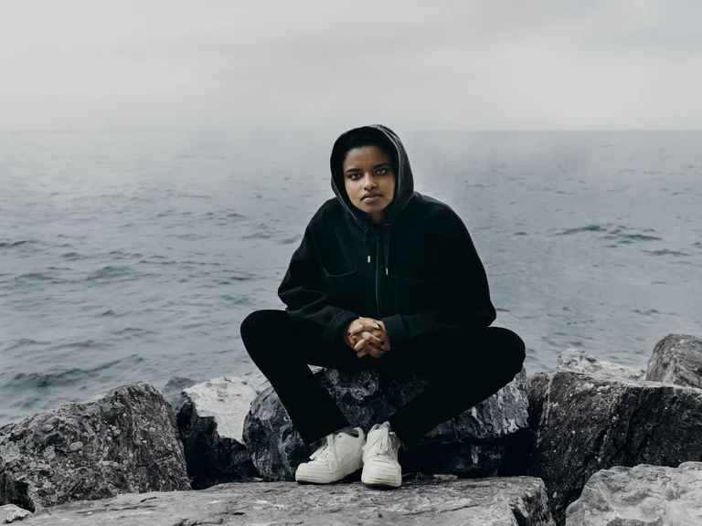 Meskerem Mees sitzt auf großen Steinen am Ufer, trägt einen schwarzen Hoodie und schwarze Kleidung und blickt direkt in die Kamera. Hinter Meskerem Mees erstreckt sich ruhiges Wasser unter einem grauen, bewölkten Himmel.