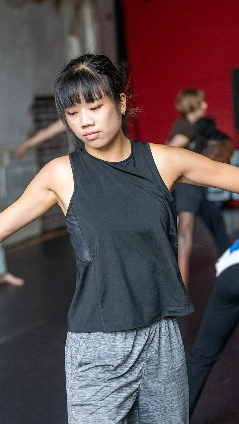 Several people rehearse contemporary dance in a studio with a red back wall; in the foreground a dancer moves with outstretched arms.