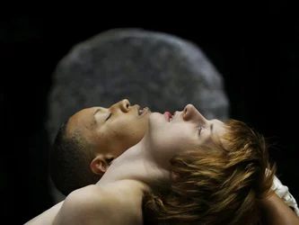 Scene from Balkan Erotic Epic: Two people lie close together with their heads tilted upward, eyes closed. The background is dark and minimal.