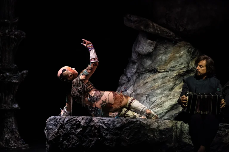A performer reclines on a rock, torso raised, with one arm expressively reaching upward. To the right, a musician with a bandoneon watches the scene attentively.