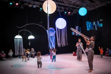 Szene aus der partizipativen Performance UNIVERS: Kinder erkunden den Raum unter schwebenden, leuchtenden Objekten, während Performer:innen Bewegungen und Klänge anleiten.