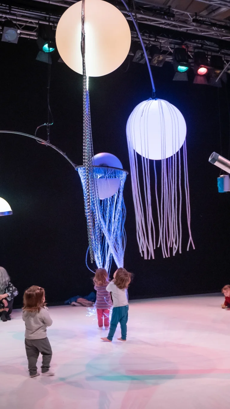 Szene aus der partizipativen Performance UNIVERS: Kinder erkunden den Raum unter schwebenden, leuchtenden Objekten, während Performer:innen Bewegungen und Klänge anleiten.