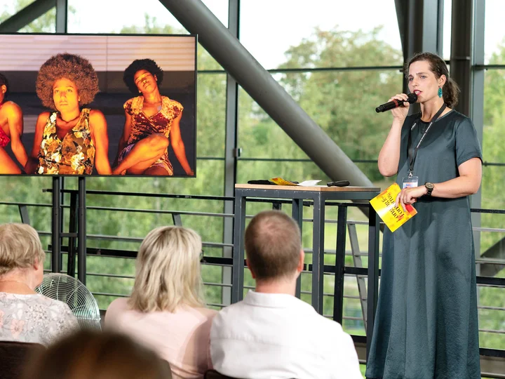 Ruhrtriennale-Dramaturgin bei ihrer Einführung zu "I Did It My Way" im Foyer der Jahrhunderthalle Bochum. Neben ihr ist ein Bildschirm platziert, der eine Einstellung aus der Produktion zeigt.