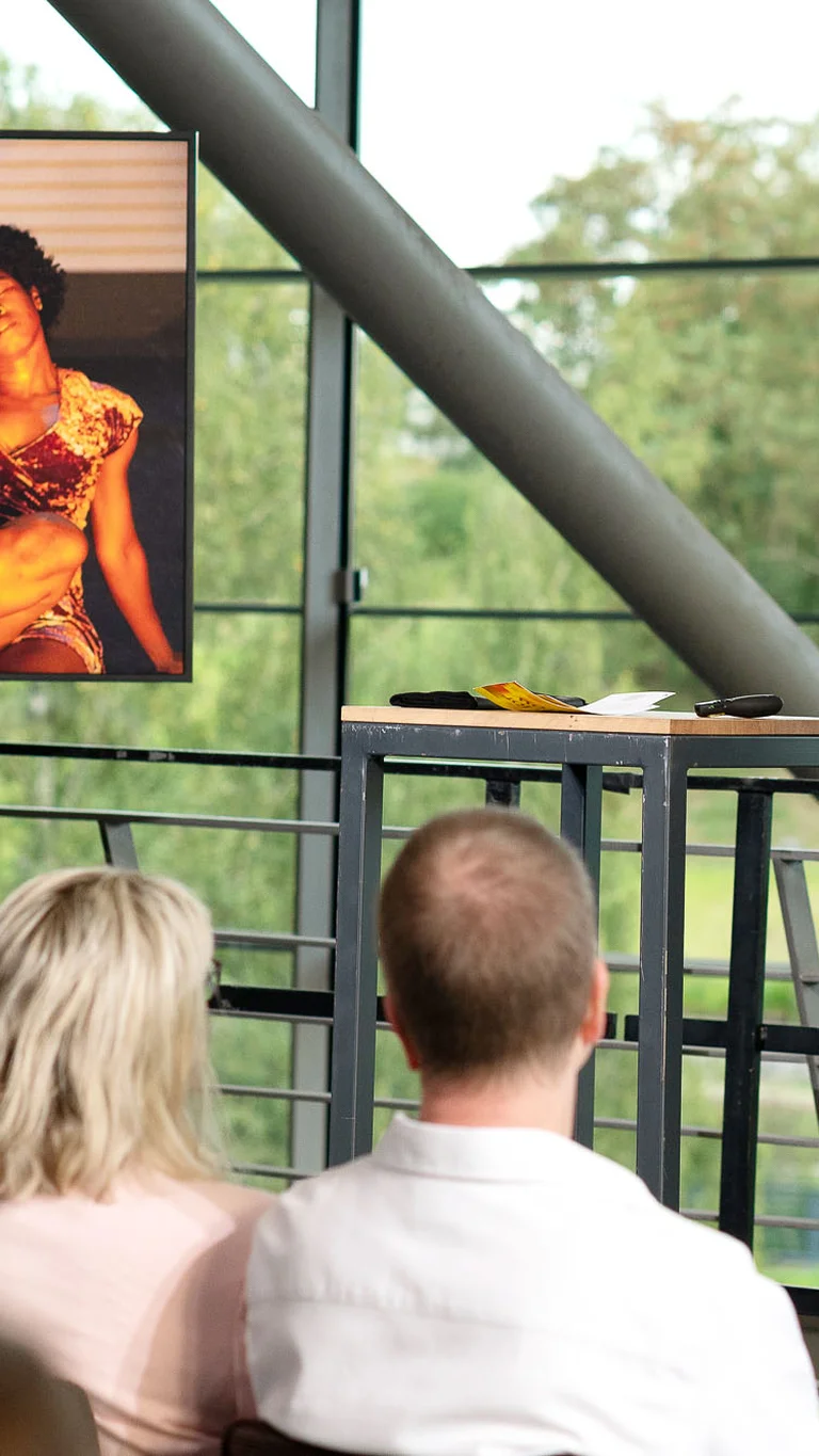 Ruhrtriennale dramaturge during her introduction to ‘I Did It My Way’ in the foyer of the Jahrhunderthalle Bochum. Next to her is a screen showing a scene from the production.