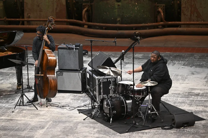 Double bassist Harish Ragshavan and drummer Tyshawn Sorey of the Tyshawn Sorey Trio perform on a stage in an industrial hall. On the left, Ragshavan stands at the double bass, while on the right Sorey sits at the drum set. Both are dressed in black, with cables, amplifiers, and part of the old turbine visible in the background.
