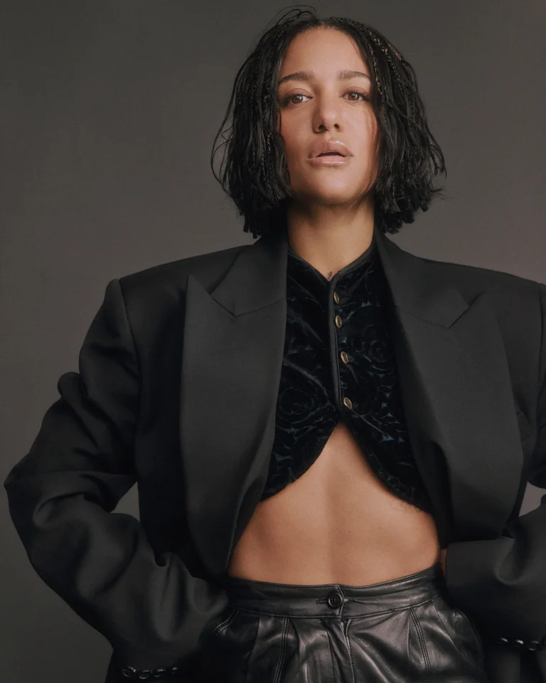 Larissa Sirah Herden trägt ein schwarzes, bauchfreies Outfit mit einem Samtoberteil, goldenen Knöpfen, einer übergroßen schwarzen Jacke und einer schwarzen Lederhose. Sie steht vor einem neutralen Hintergrund.