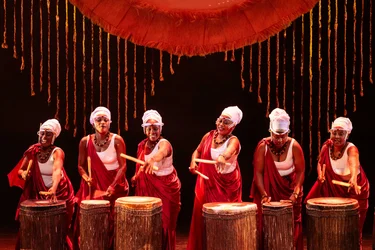 Sechs Performerinnen schlagen auf große Trommeln. Rotes Licht und hängende Schnüre rahmen die Szene.