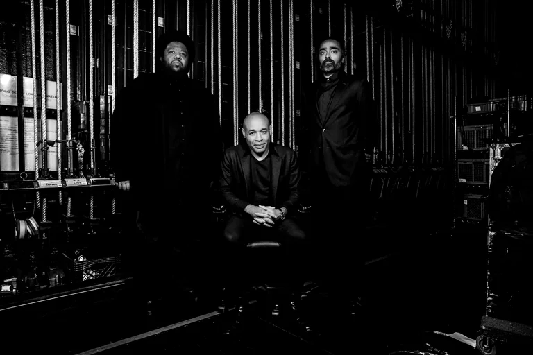 Das Trio aus Tyshawn Sorey, Aaron Diehl und Harish Raghavan ist dunkel gekleidet und blickt in die Kamera. Tyshawn Sorey und Harish Raghavan stehen, Aaron Diehl sitzt zwischen ihnen. Es handelt sich um ein schwarz-weiß Foto.