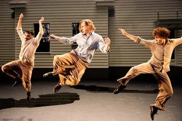 Lars Eidinger and two men jump simultaneously on stage with expressive body posture — their movements are powerful and synchronized.