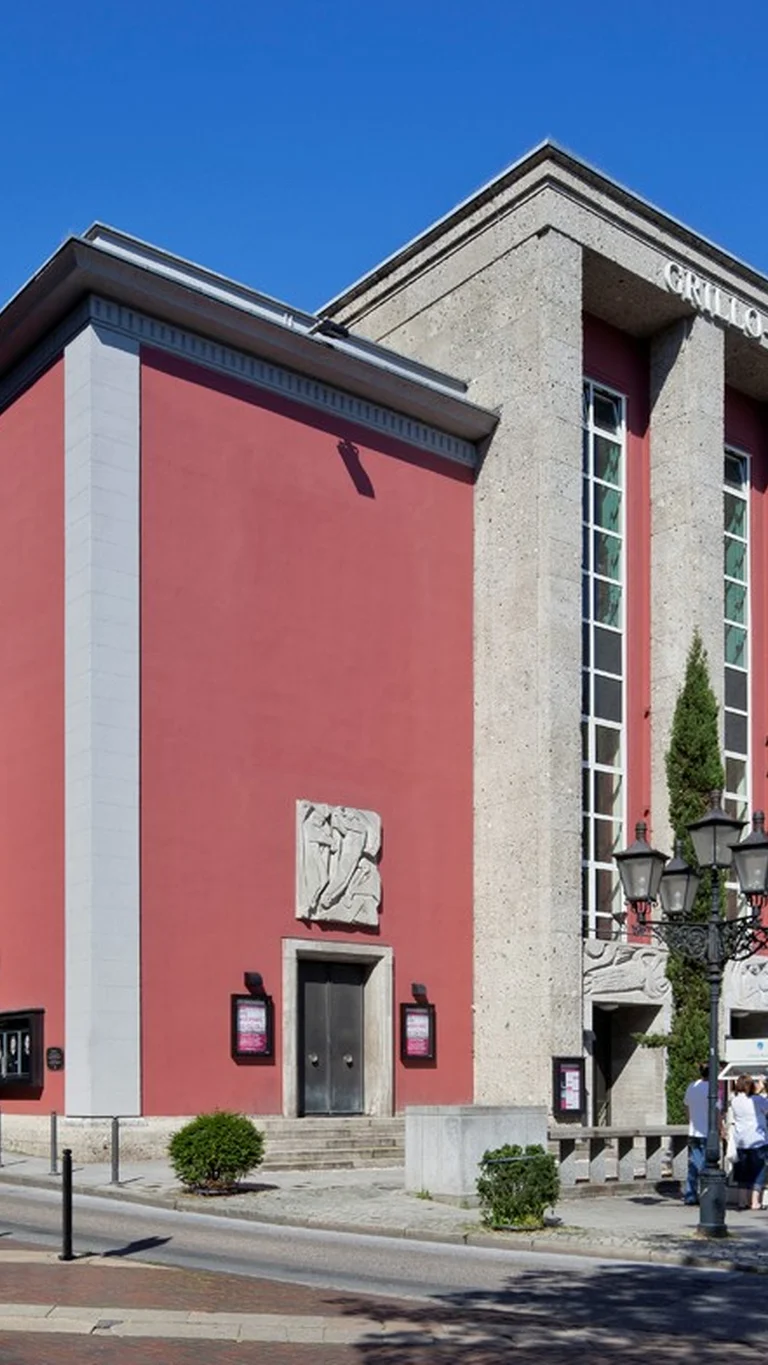 Außenansicht des Grillo-Theaters in Essen mit roter Fassade, hohen steinernen Pfeilern am Eingangsbereich und Vorplatz mit Fahrrädern und Passant:innen bei Tageslicht.