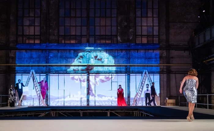 Several performers stand on an elevated stage in front of the large window facade of a former industrial hall. In the center of the image, a projection of a nuclear mushroom cloud creates a dramatic atmosphere. On the right side of the image, a woman in a silver sequin dress walks toward the stage, while other performers are positioned between ladders and white panels.