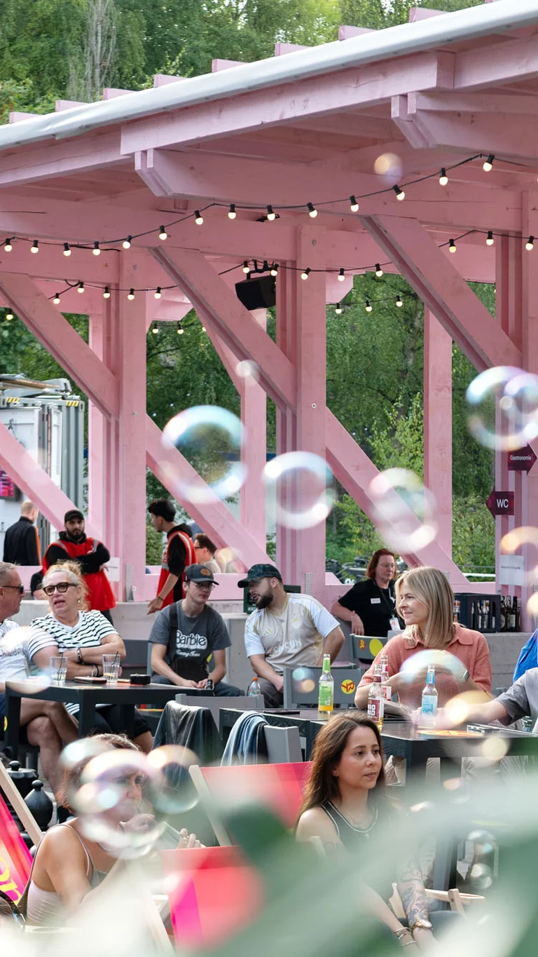 Besucher:innen einer Open-Air-Veranstaltung sitzen und stehen unter der rosafarbenen Holzkonstruktion des Festivalzentrums Wunderland; im Vordergrund sind schwebende Seifenblasen zu sehen.