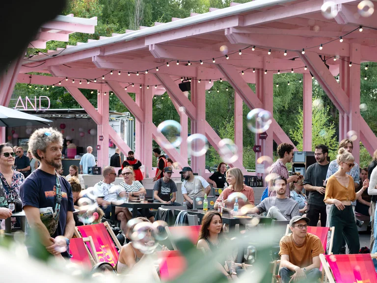 Besucher:innen einer Open-Air-Veranstaltung sitzen und stehen unter der rosafarbenen Holzkonstruktion des Festivalzentrums Wunderland; im Vordergrund sind schwebende Seifenblasen zu sehen.
