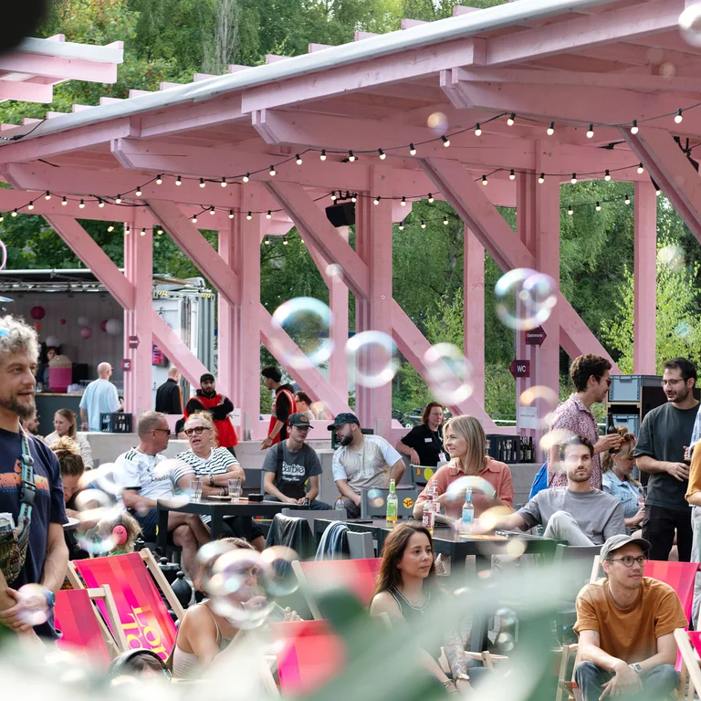 Besucher:innen einer Open-Air-Veranstaltung sitzen und stehen unter der rosafarbenen Holzkonstruktion des Festivalzentrums Wunderland; im Vordergrund sind schwebende Seifenblasen zu sehen.
