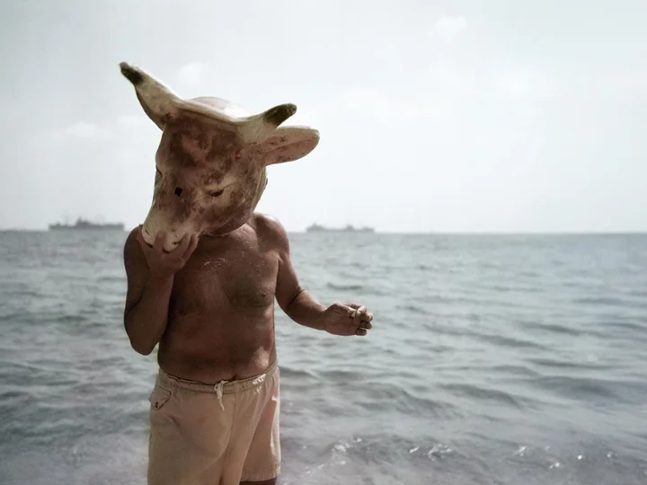 Pablo Picasso stands shirtless on a beach with the sea in the background, wearing a large, weathered bull’s head mask. The scene appears surreal and mysterious.