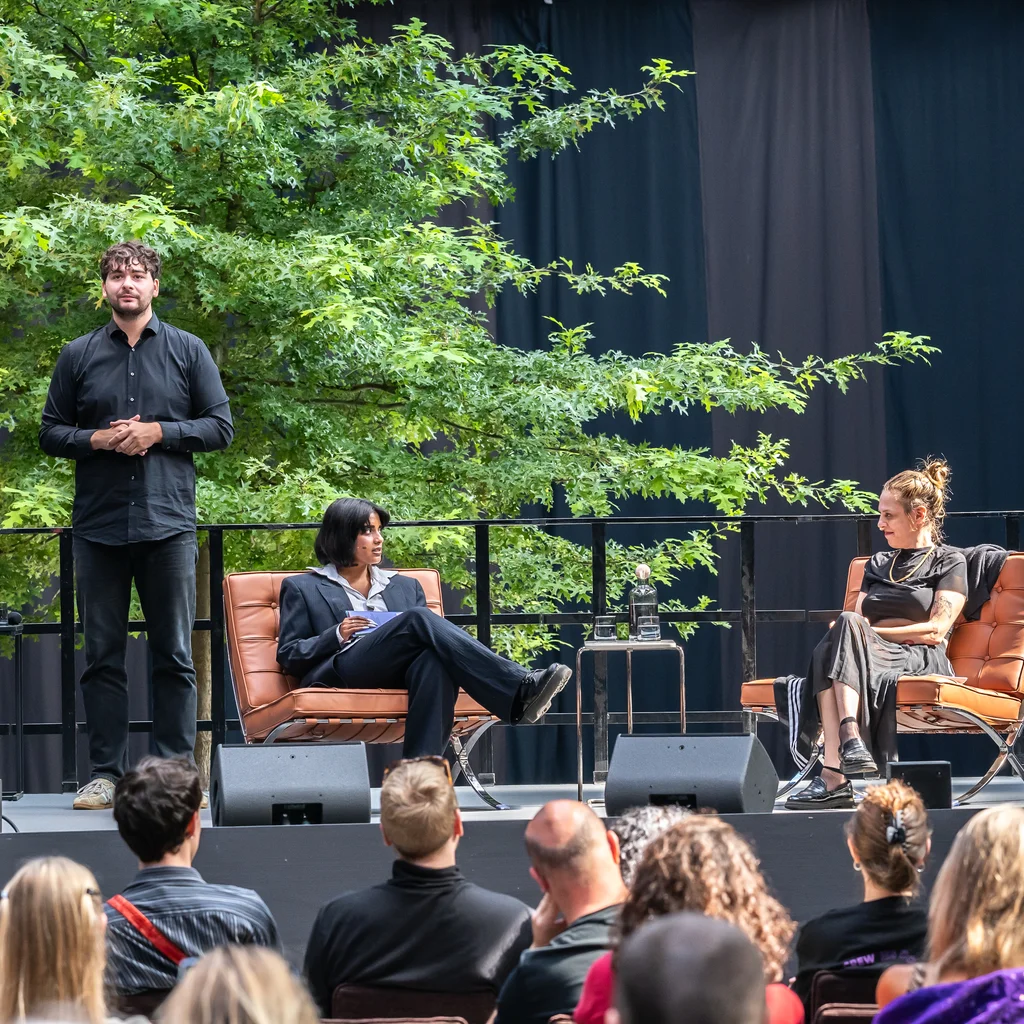A panel discussion on a stage with four people. A man in a black shirt and dark pants stands and translates into German Sign Language. Fatima Khan and Sivan Ben Yishai sit in armchairs and talk to each other.