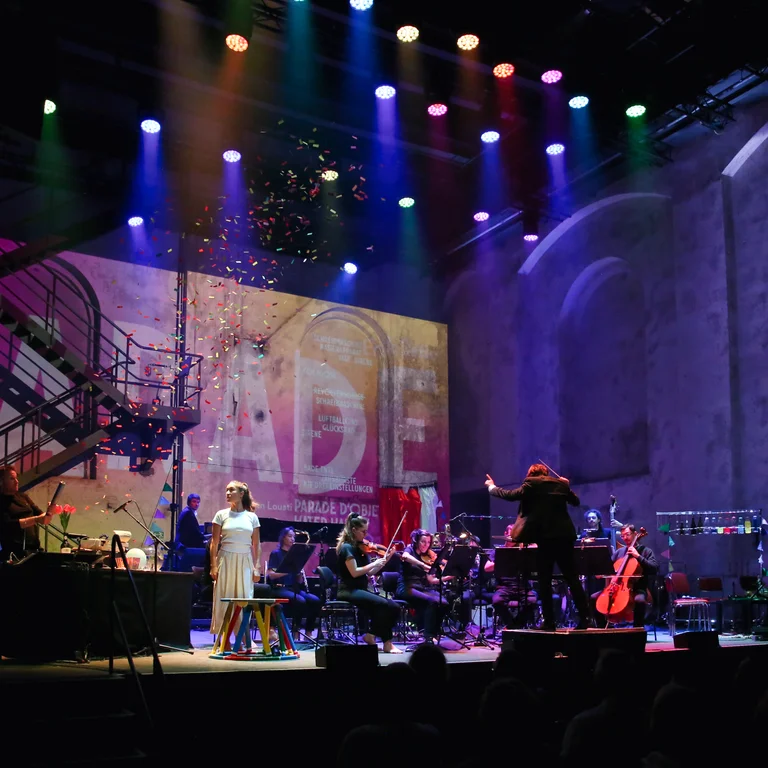 The performers at the ‘Vorhang!’ concert on stage at the Gebläsehalle in the Duisburg-Nord Landscape Park. The stage is lit up in blue, and in the background the word ‘Parade’ can be seen on the wall.