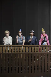 Four performers stand behind a wooden balustrade. Two wear colorful party hats and one wears a pink flower garland. They hold glasses of water and look ahead.