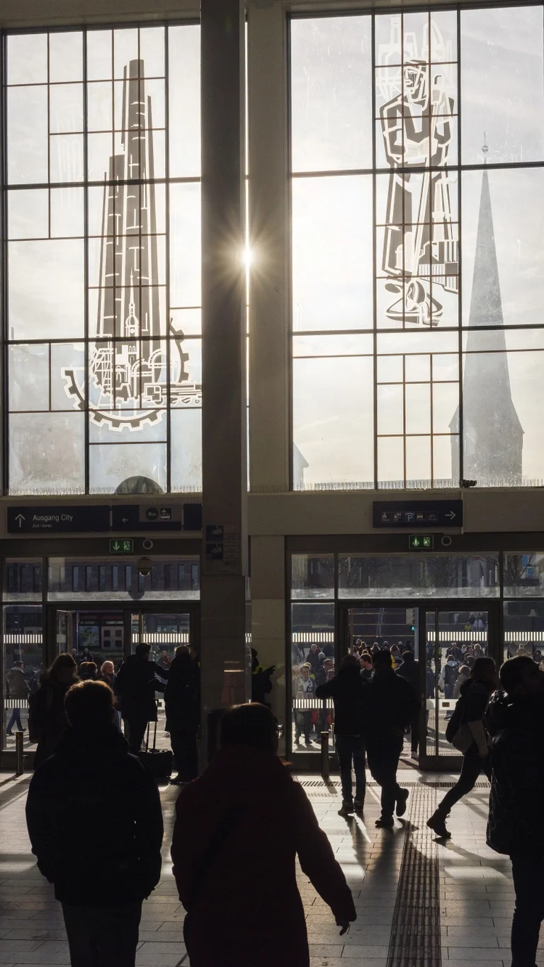 Station hall in Dortmund with a glass façade and sunlight streaming in; several people standing or walking inside.
