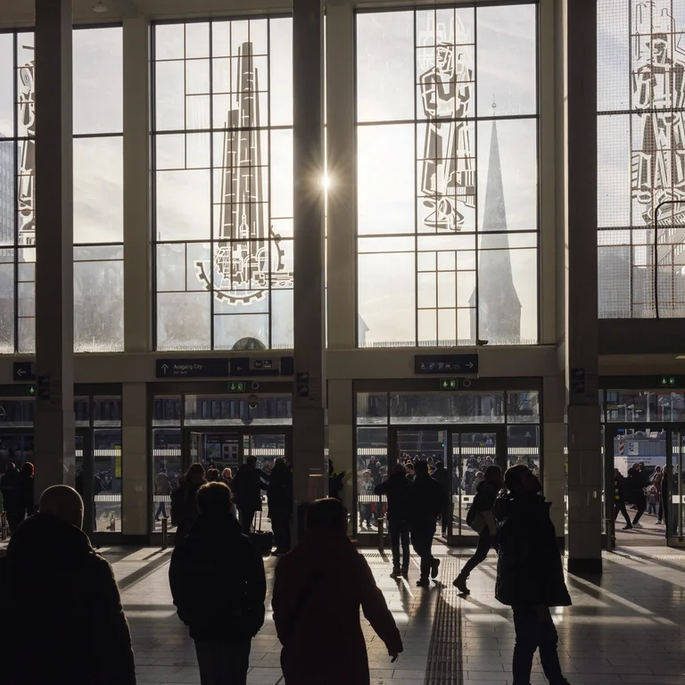 Bahnhofshalle in Dortmund mit Glasfassade und einfallendem Sonnenlicht; mehrere Personen stehen oder gehen im Innenraum.
