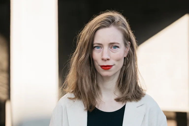 Eine junge Frau mit offenem Haar. Sie trägt ein offenes Hemd über einem schwarzen Oberteil und blickt direkt in die Kamera.