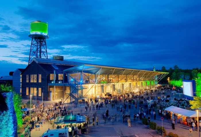 Die Jahrhunderthalle Bochum bei Nacht, beleuchtet mit einem grün angestrahlten Wasserturm und zahlreichen Besucher:innen auf dem Vorplatz.