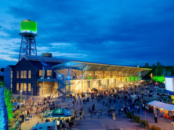 Die Jahrhunderthalle Bochum bei Nacht, beleuchtet mit einem grün angestrahlten Wasserturm und zahlreichen Besucher:innen auf dem Vorplatz.