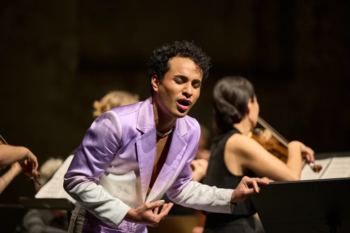 Samuel Mariño im lilafarbenen Anzug singt ausdrucksstark, begleitet von einer Violinistin im Hintergrund.