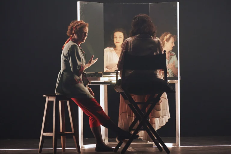 Two actors at a dressing table, one seated in front of the mirror, the other seen talking at the side.