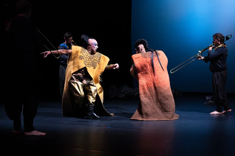 Two performers in colorful costumes dance on stage while musicians with trombones play in the background. The scene combines expressive movement with music.