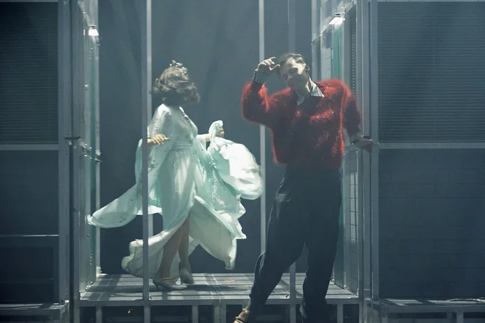 Two actors in motion on stage – one wearing a red sweater, the other in a flowing light blue costume with a crown.