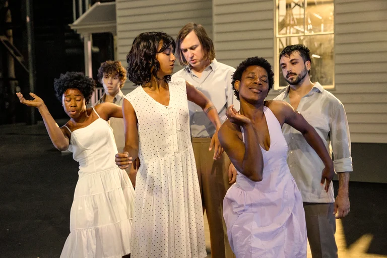 Six performers stand on a theatre stage in front of a light-colored house facade. In the foreground, three women in white dresses perform expressive dance movements. In the center, Larissa Sirah Herden and Lars Eidinger face each other. Two additional performers stand in the background, observing the scene.