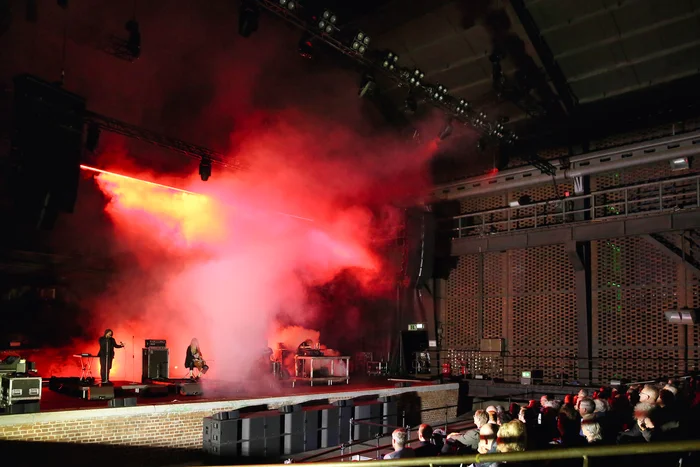 Live concert with red light and smoke, musicians on stage, audience in the foreground.