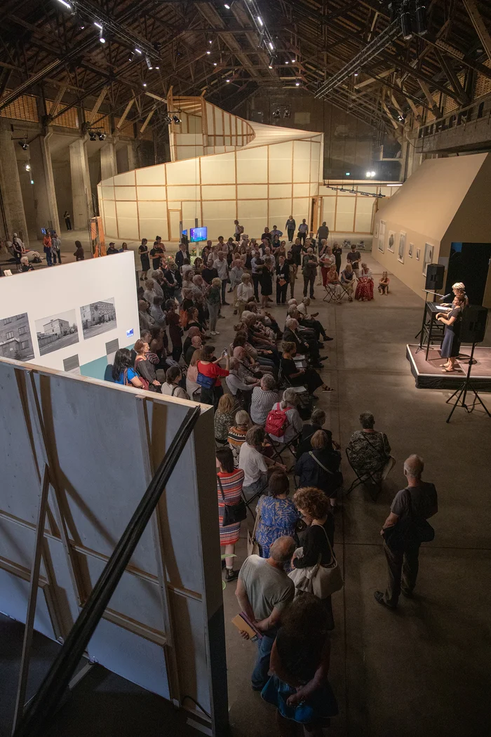 Das Bild zeigt die Ausstellungseröffnung „Landscapes of an Ongoing Past“ von Urbane Künste Ruhr im Rahmen der Ruhrtriennale 2024. Die Veranstaltung findet im Salzlager der Kokerei Zollverein in Essen statt. Eine große Menschenmenge hat sich versammelt, um den Vorträgen und Präsentationen zu lauschen. Im Hintergrund sind Teile der Ausstellung sowie die eindrucksvolle Architektur des Raumes zu sehen. Die Atmosphäre ist lebendig und interessiert, während die Besucher:innen die Kunstwerke betrachten und den Redner:innen zuhören.