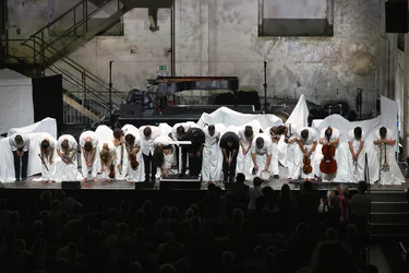 The musicians of the ensemble Les Apaches ! bow together at the front of the stage before the audience. They are dressed predominantly in white, with some holding their instruments. In the background, a grand piano and stage elements covered with white cloths stand against the walls of the industrial hall.