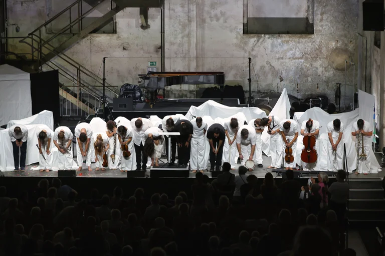The musicians of the ensemble Les Apaches ! bow together at the front of the stage before the audience. They are dressed predominantly in white, with some holding their instruments. In the background, a grand piano and stage elements covered with white cloths stand against the walls of the industrial hall.