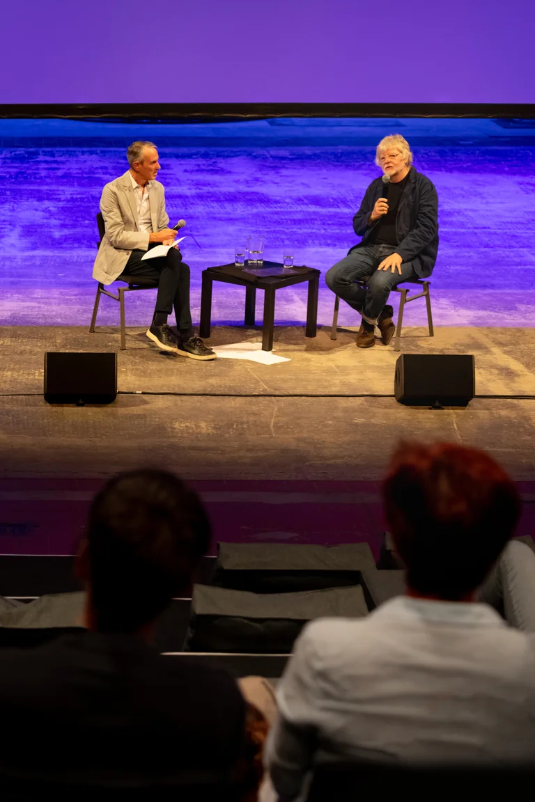 Ivo Van Hove and Will Gregory sitting on stage with microphones, speaking while the audience listens.