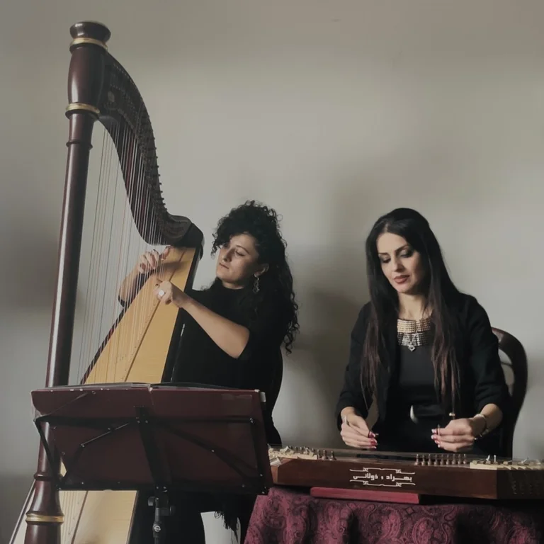 The musicians Zainab Lax and Areezo Molaei are playing a large concert harp and a santur (a Persian hammered dulcimer). Both are wearing black clothing. In front of them stands a music stand and a table covered with a cloth.