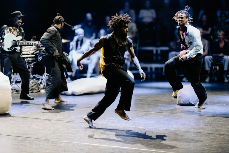 Three dancers are on stage. The dancer in the foreground jumps up with his right leg. In the background, a musician wearing a hat stands playing the guitar.