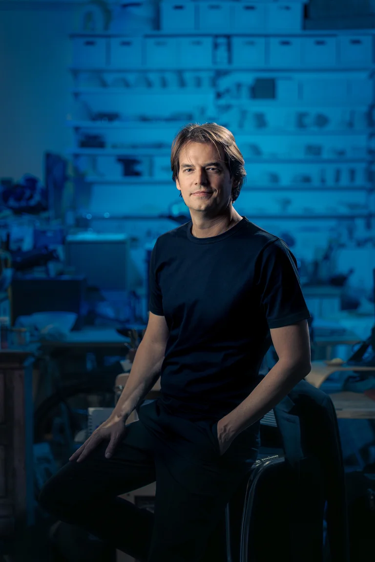 Julien Masmondet stands in a black T-shirt stands in a blue-lit studio in front of shelves with work materials.