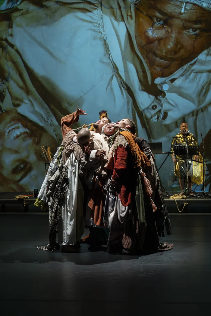 Mehrere Performer:innen in bunten Gewändern stehen eng umschlungen auf der Bühne, im Hintergrund ist eine große Projektion von Gesichtern zu sehen.